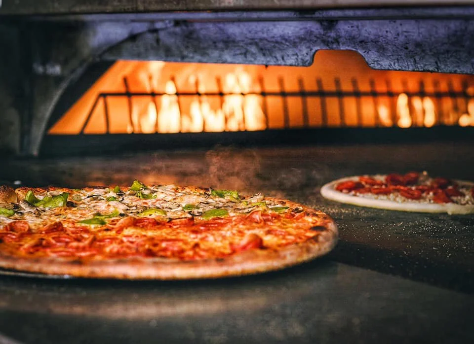 Multiple pizzas baking inside Amici’s brick oven with visible flames and stone cooking surface.