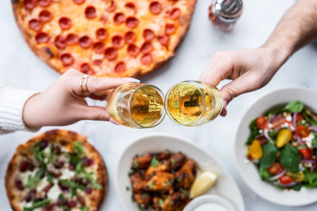 A couple cheering their champagne glasses with an Amici’s pizza and sides in the background.