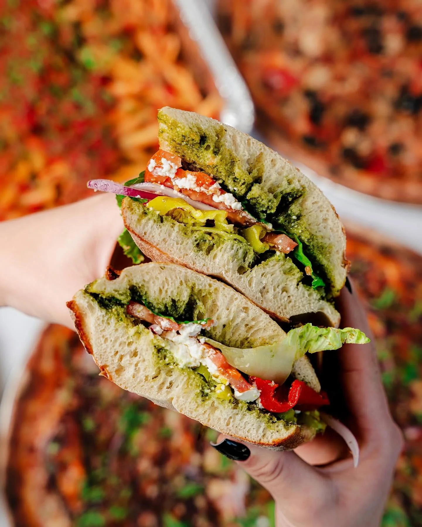 Hands holding Amici’s vegetarian Greek Sandwich with other vegetarian pizza and sides in the background.
