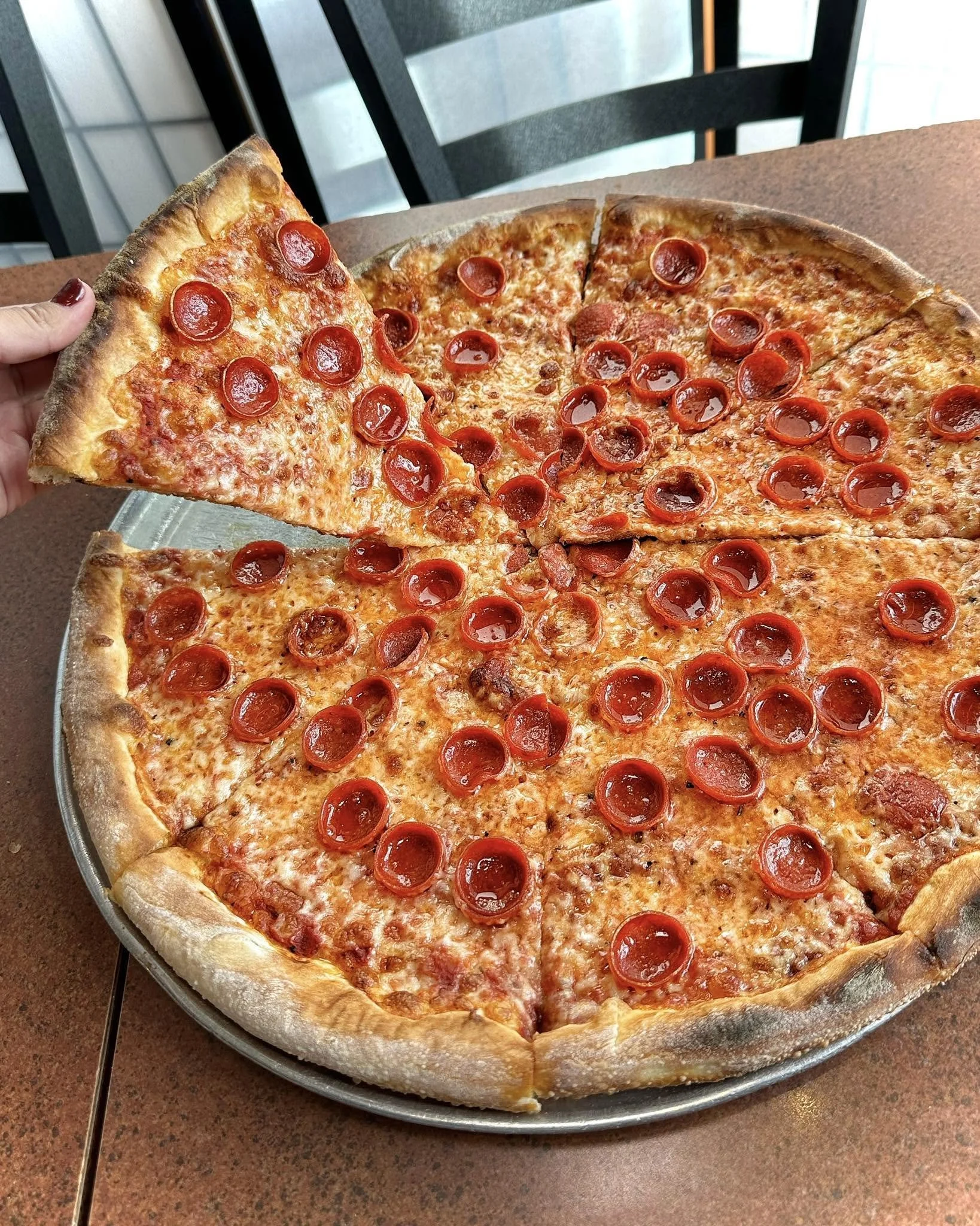 Hand lifting a slice of Amici’s New York-style pepperoni pizza, showing its thin, chewy, foldable crust.
