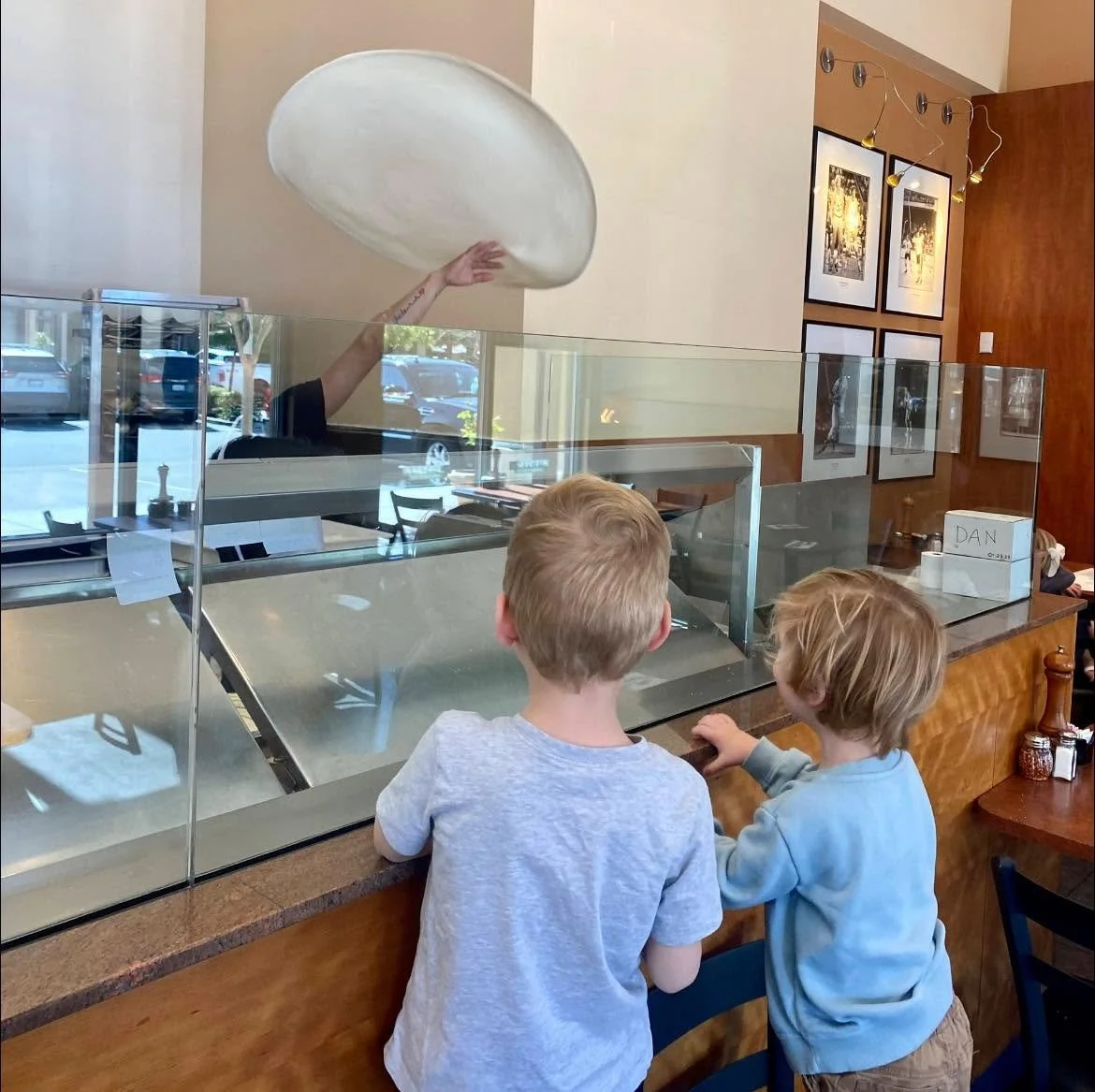 Two young boys watch in amazement as an Amici's chef tosses fresh pizza dough high into the air during pizza preparation.