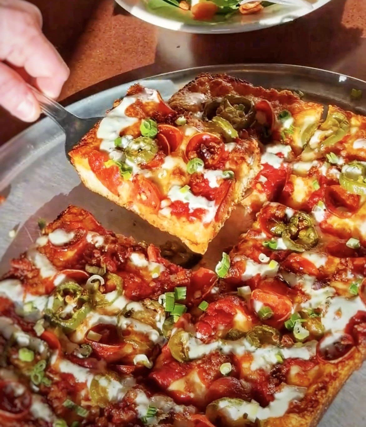 A hand lifts a thick slice of Detroit-style pizza from Amici’s with pepperoni, jalapenos, and chopped green onions.