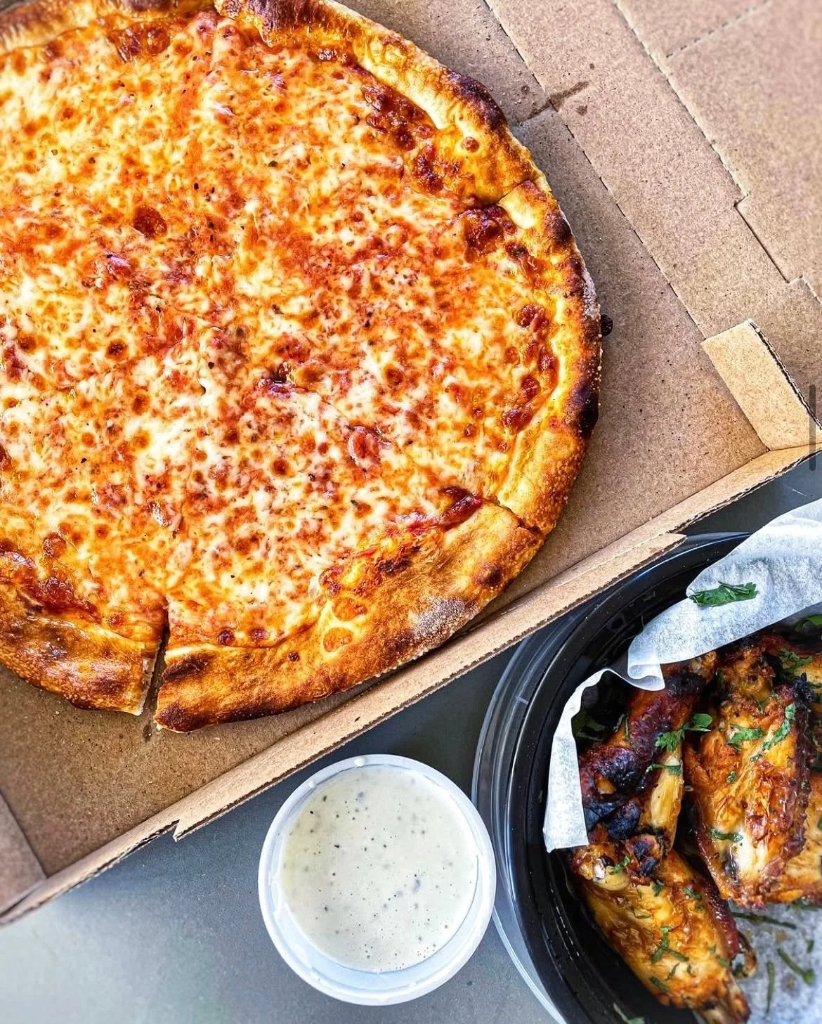 Fresh Amici’s cheese pizza in an open box served alongside a bowl of roasted chicken wings for a complete meal.