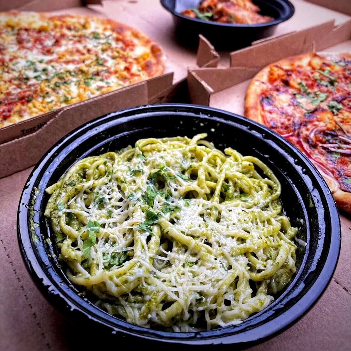 Amici’s pesto pasta in the foreground with two pizzas (one white, one red) and a side of baked pasta behind it.