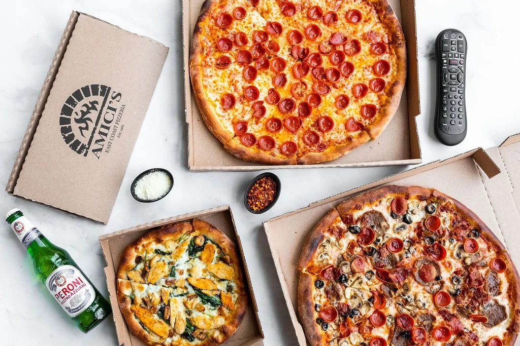 Three different-sized pizzas from Amici’s in takeout boxes, along with an Italian side and a wine bottle on a table.