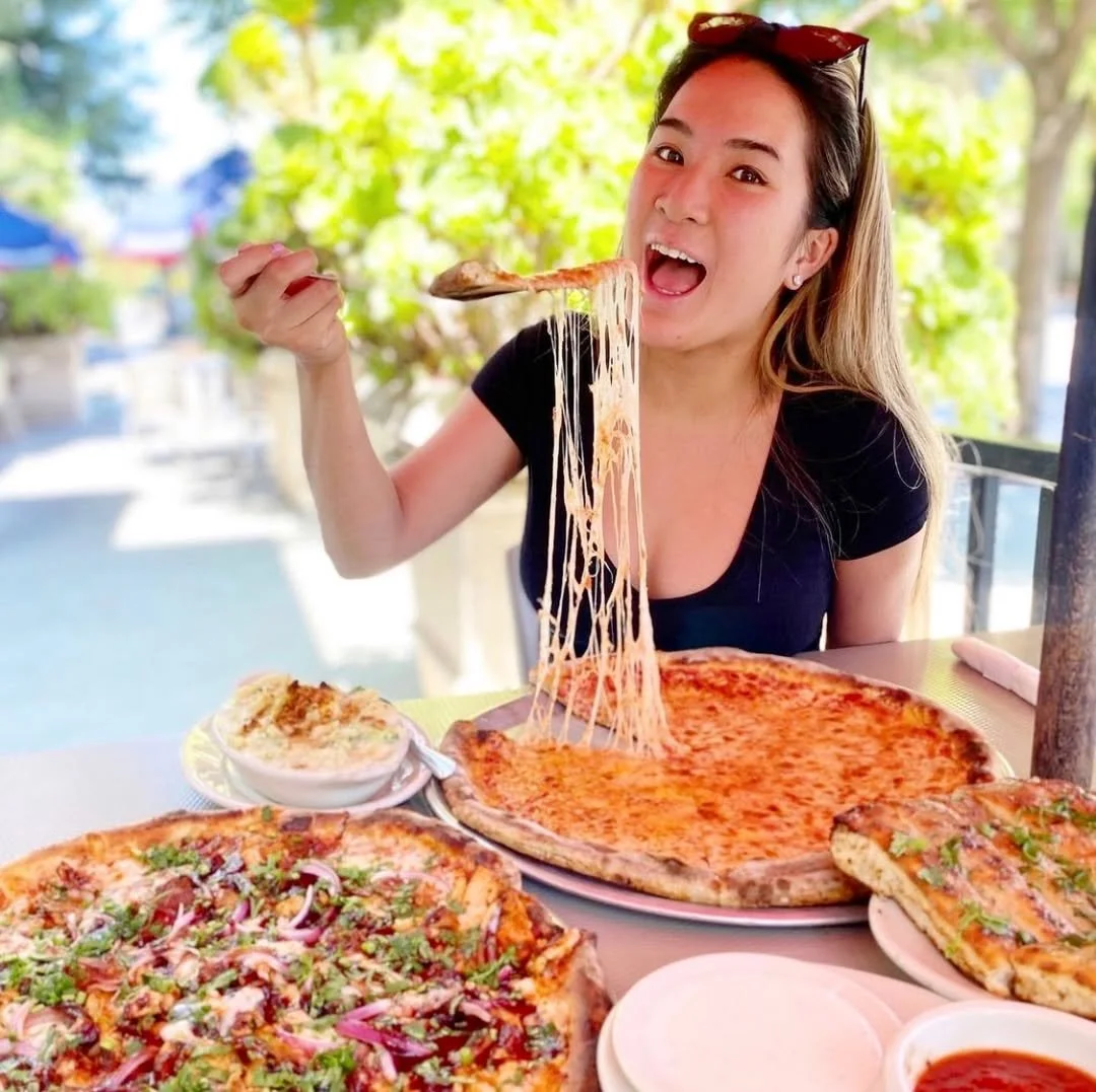 A woman eats a New York-style cheese pizza from Amici’s with a fork at an outdoor table with pizza and side dishes.