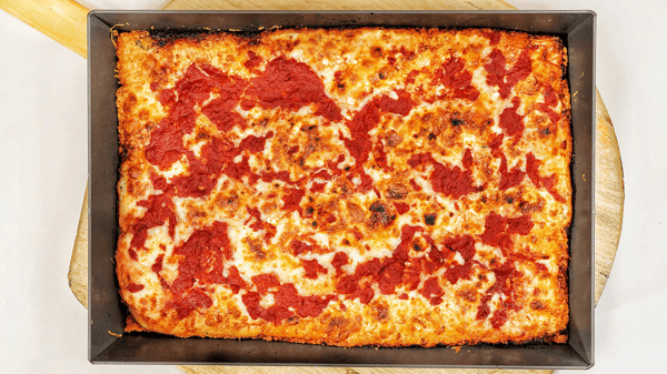 Amici's Three Cheese Red Top Detroit-style pizza with caramelized cheese edges served in a rectangular pan on a wooden board.