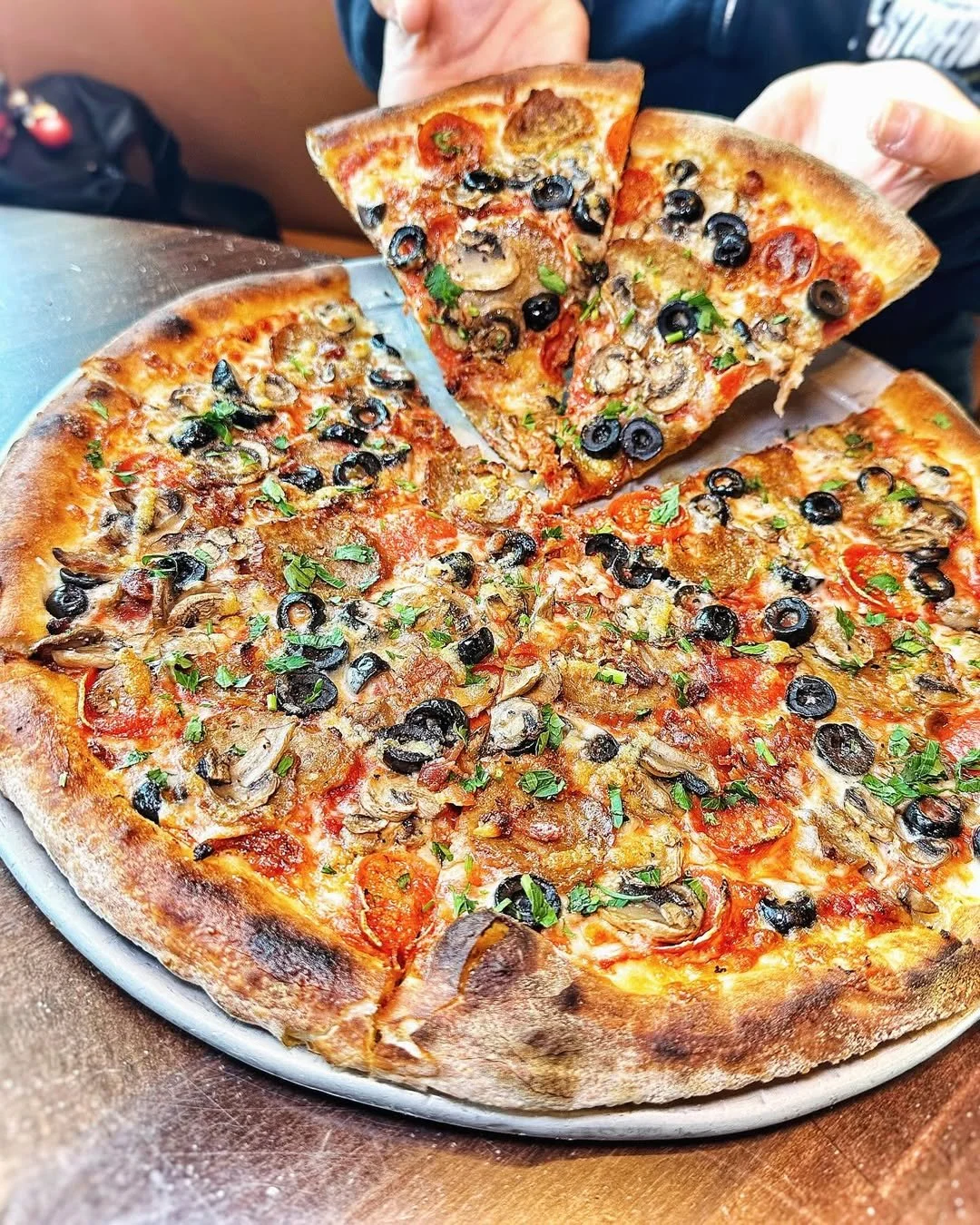 A gluten-free veggie pizza from Amici’s on a tray with 2 slices being pulled up by a person.