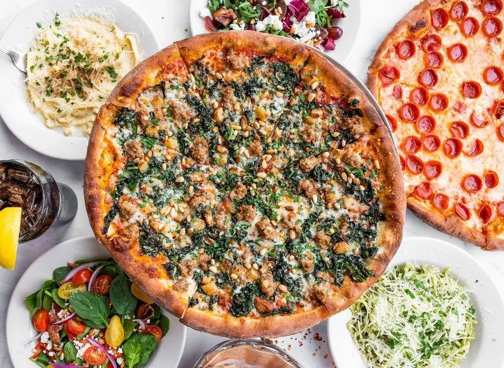 Overhead view of an Amici's kale and sausage pizza surrounded by salads and pasta dishes.
