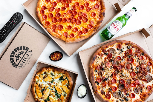 Overhead view of three Amici’s pizzas in boxes, with beer, parmesan, chili flakes, and a TV remote.
