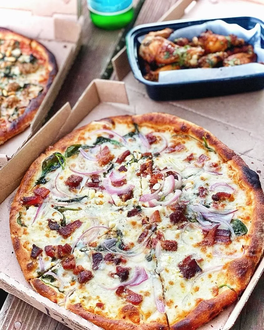 A person cutting into a pizza topped with spinach, onions, pancetta, herbs, and mozzarella.