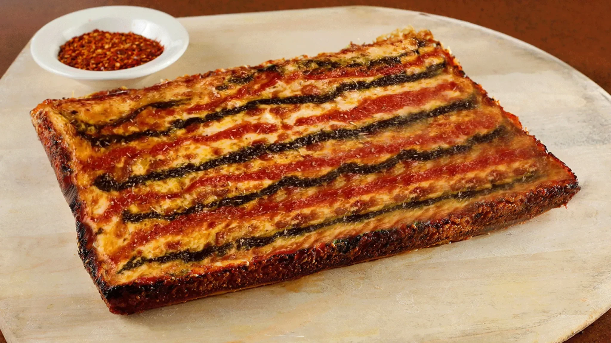 Amici's MVP Detroit-style pizza topped with signature sauces beside a bowl of chili flakes on a wooden serving board.
