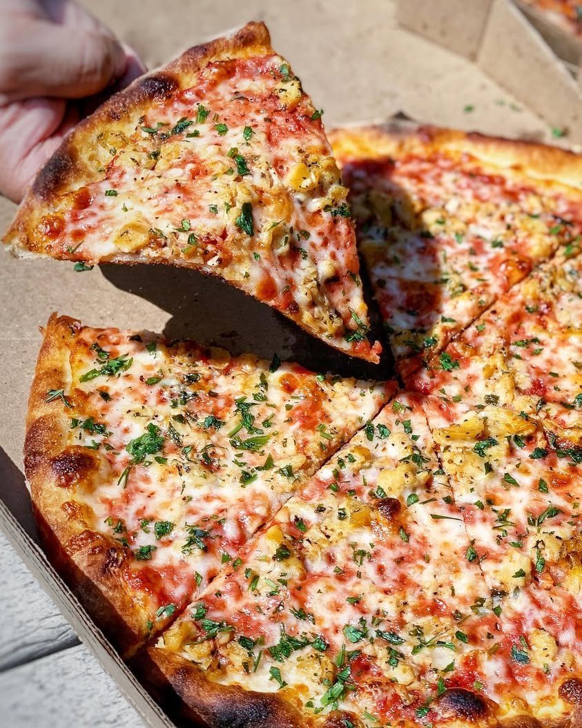 Hand lifting a slice of Amici’s Manhattan Red Clam New York-style pizza from a takeout box, showing its thin, foldable crust.