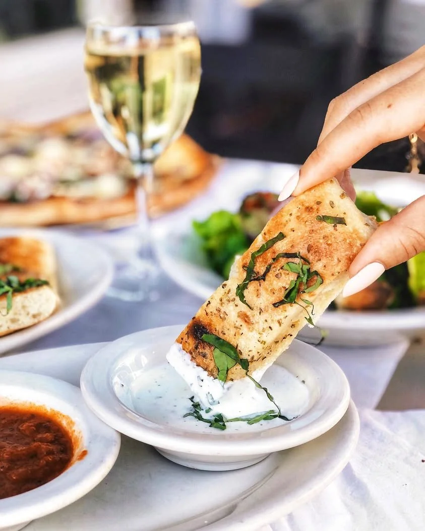 A hand dipping a garlic breadstick in a bowl of cheese with pizza, champagne, and other dishes on the table at Amici’s.