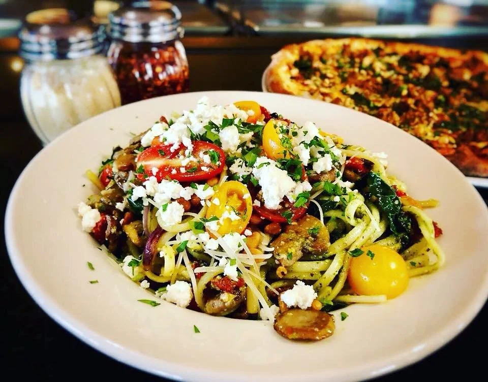 Close-up of a vegetarian pasta dish from Amici’s with fresh tomatoes and feta cheese, with pizza in the background.