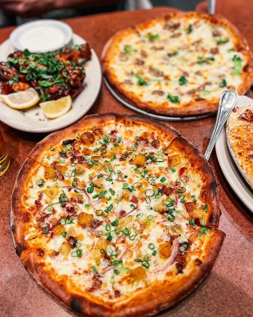 Amici’s pizza spread: NY slices, chicken wings, and a flatbread on a table, with a potato pizza in the foreground.