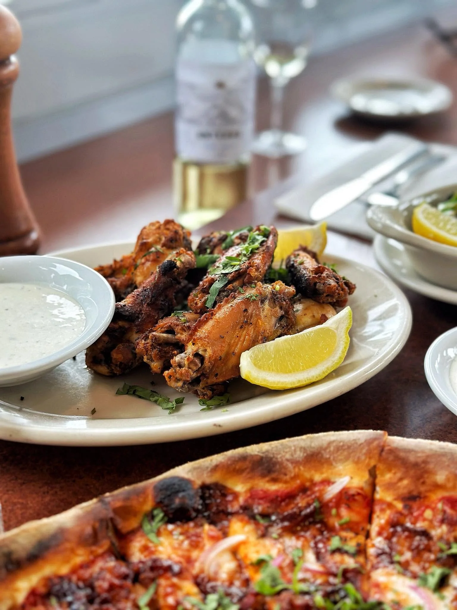 Plate of Amici's roasted chicken wings served with lemon wedges and dipping sauce beside a freshly baked pizza.