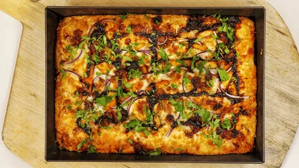 Overhead view of a rectangular Amici’s BBQ chicken pizza in a baking pan on a wooden serving board.