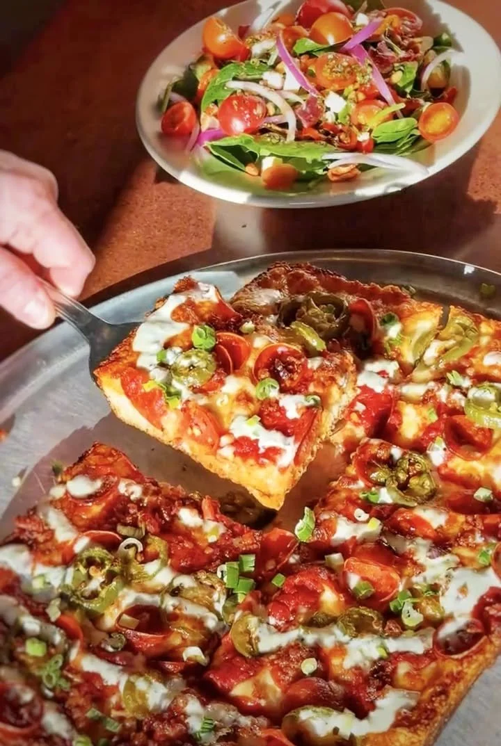 Hand pulling a square slice of Amici’s Firehouse Special Detroit-style pizza from a rectangular pan, highlighting its thick crust and crispy cheese edges.