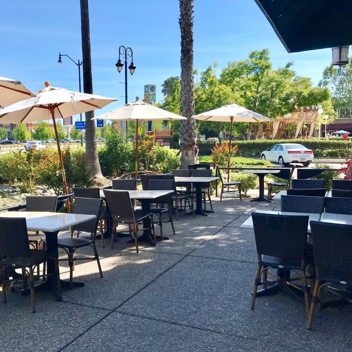 Outdoor patio at Amici's Vacaville featuring white umbrellas, palm trees, and a nearby carousel on a sunny day.
