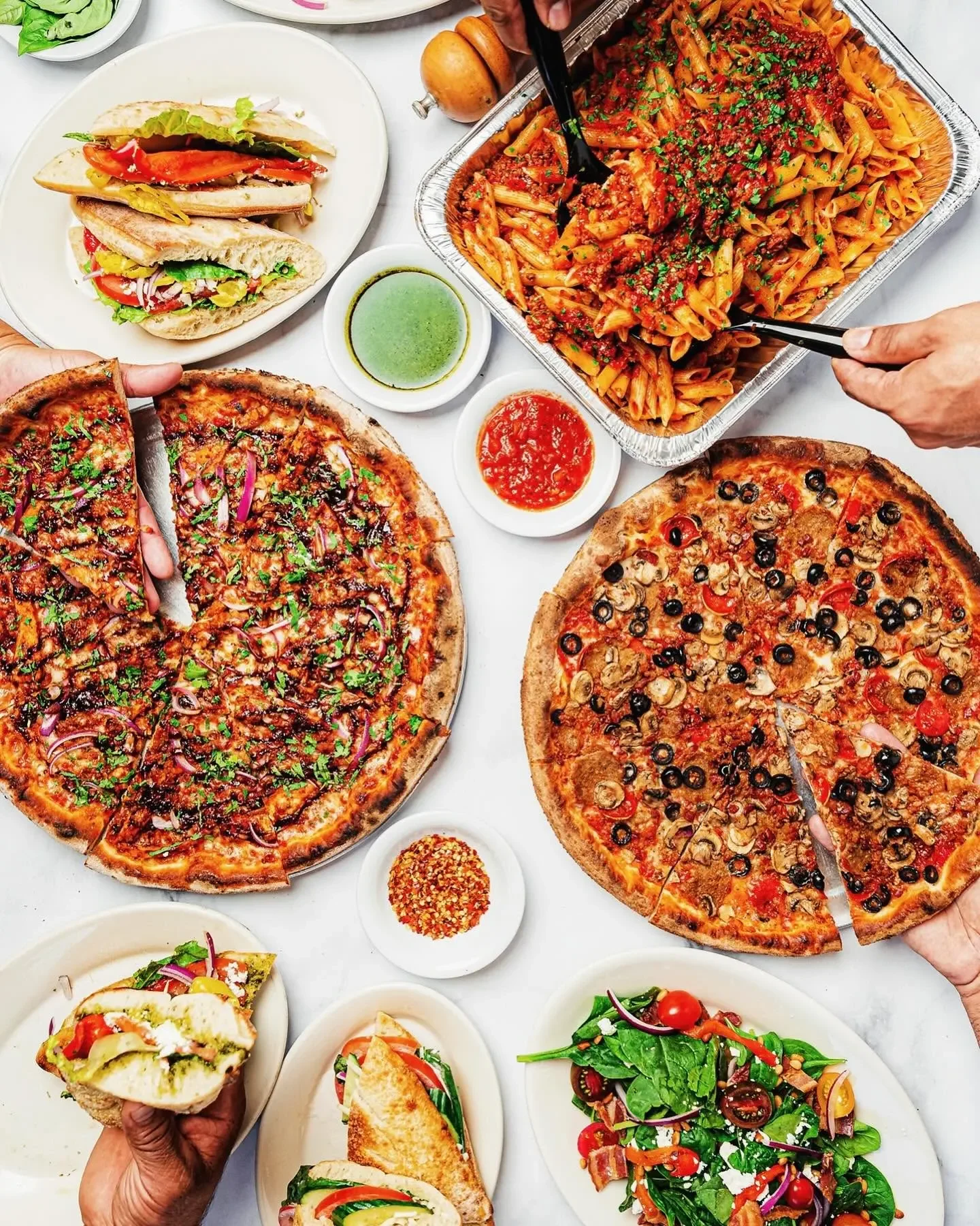 Overhead view of a large meal spread featuring Amici's pizzas, penne pasta, sandwiches, and salads.