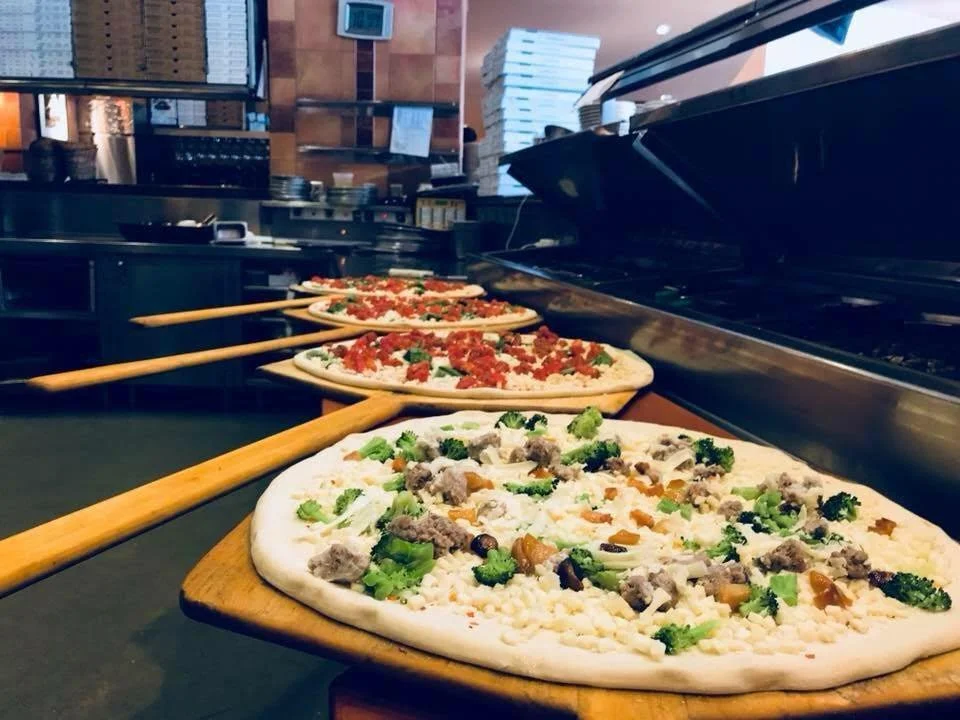 Multiple pizzas being assembled with fresh toppings in Amici’s East Coast Pizzeria kitchen.