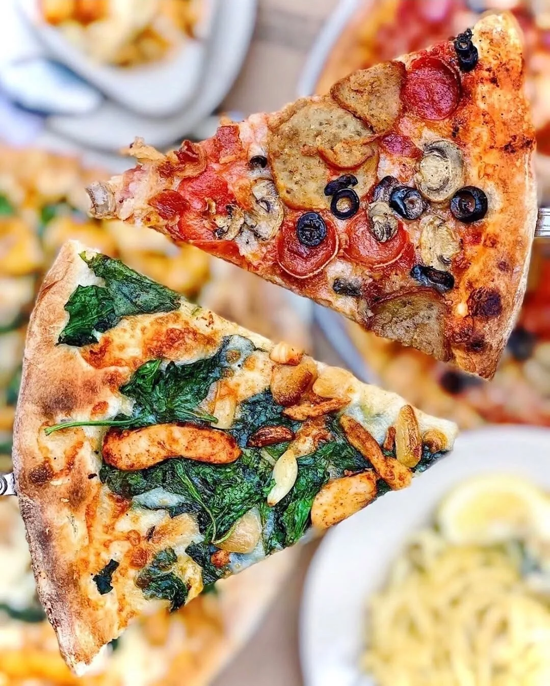 Hands holding slices of Amici's Combo and Pollo pizzas with Italian dishes and drinks on the table.