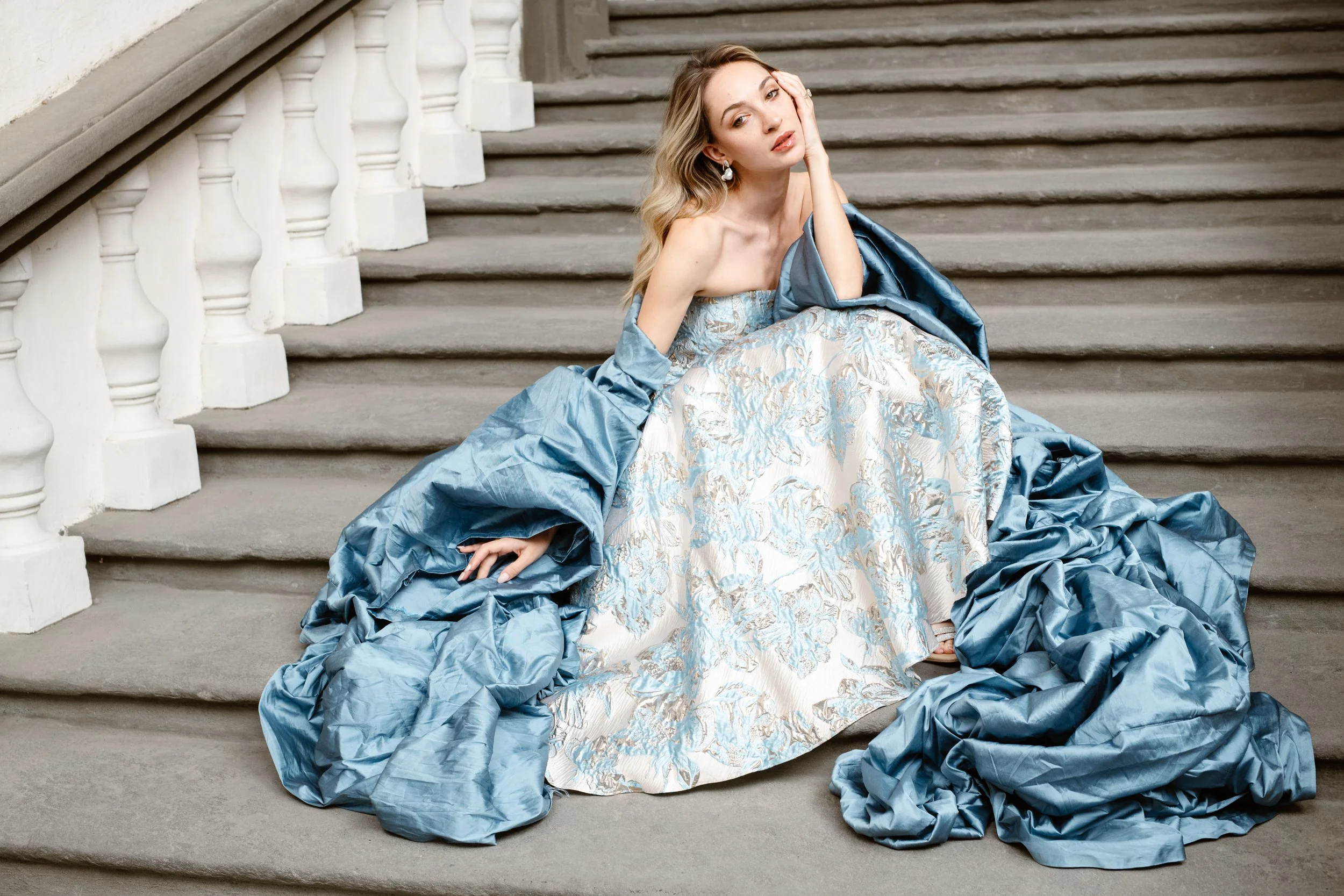 Gorgeous Raphaelite bride glows in the late afternoon light. She sits on the stairs entering an Italian garden in Lucca, Tuscany. Her designer gown shimmers, surrounded by the voluminous folds of a blue wrap.