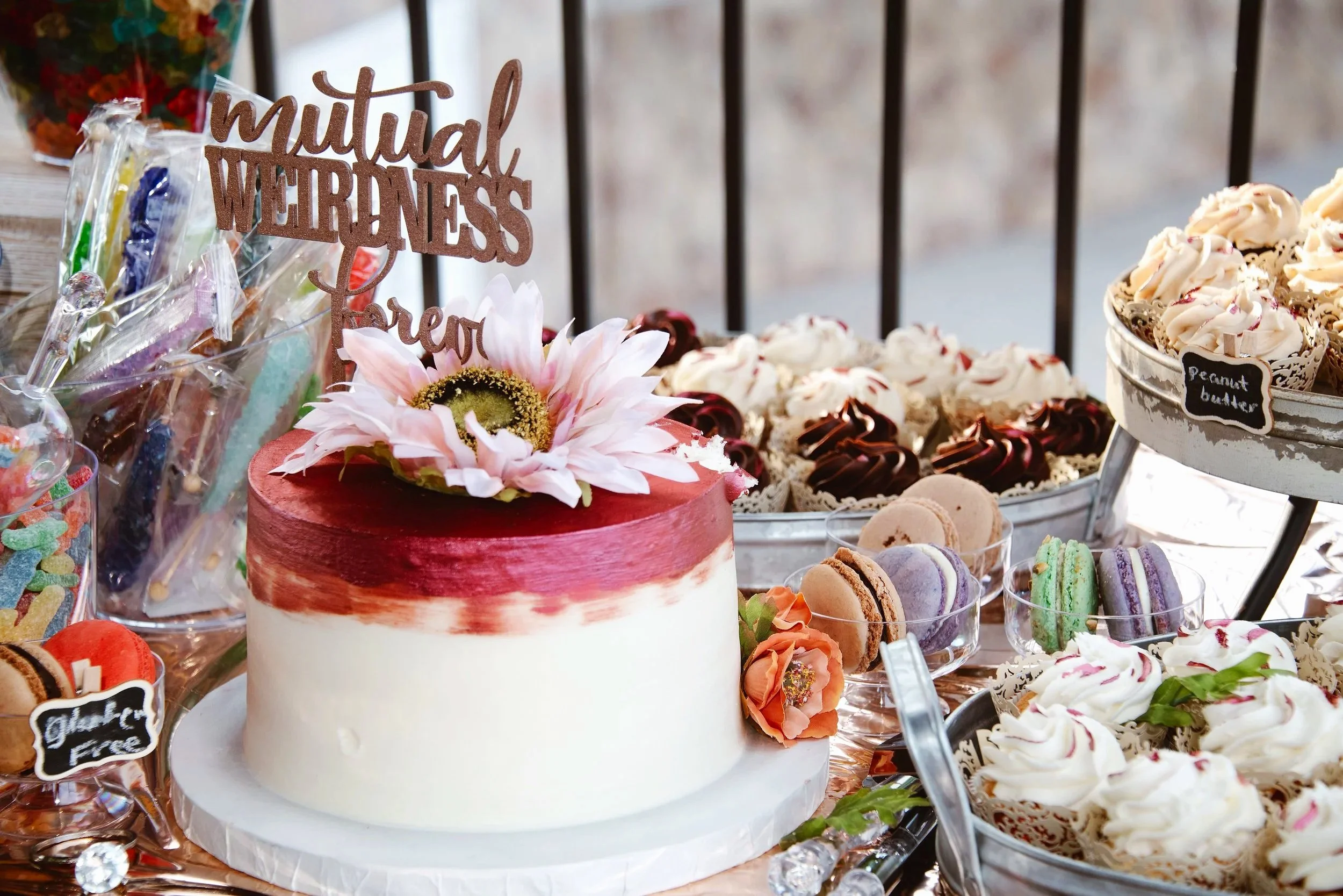 Wedding cake that has a topper saying "mutual weirdness forever." Surrounded by jars of rock candy lollipops, maccaroons, and packets of gummy worms. There is also a sign that offers gluten free macaroons.