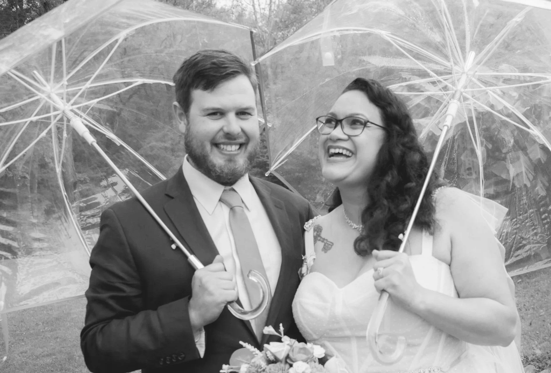 Just married couple, grinning under clear umbrellas on their wedding day. Black and white photo.
