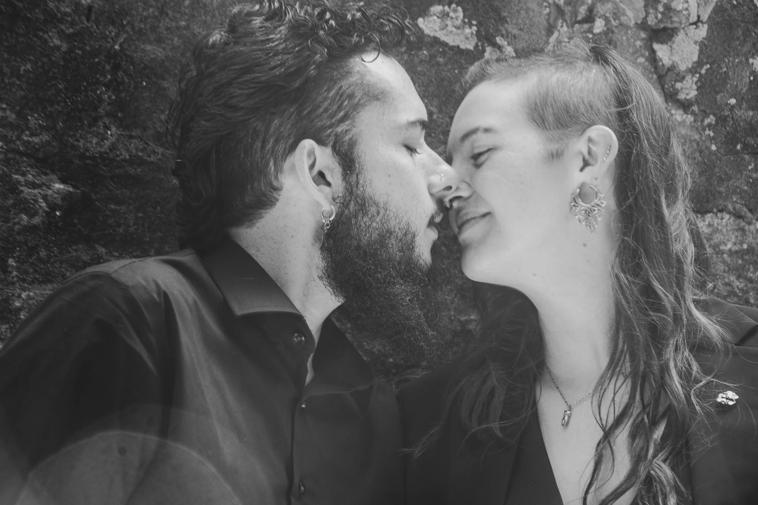 Couple about to kiss, in front of a stone wall covered in lichen. Photo is in black and white.