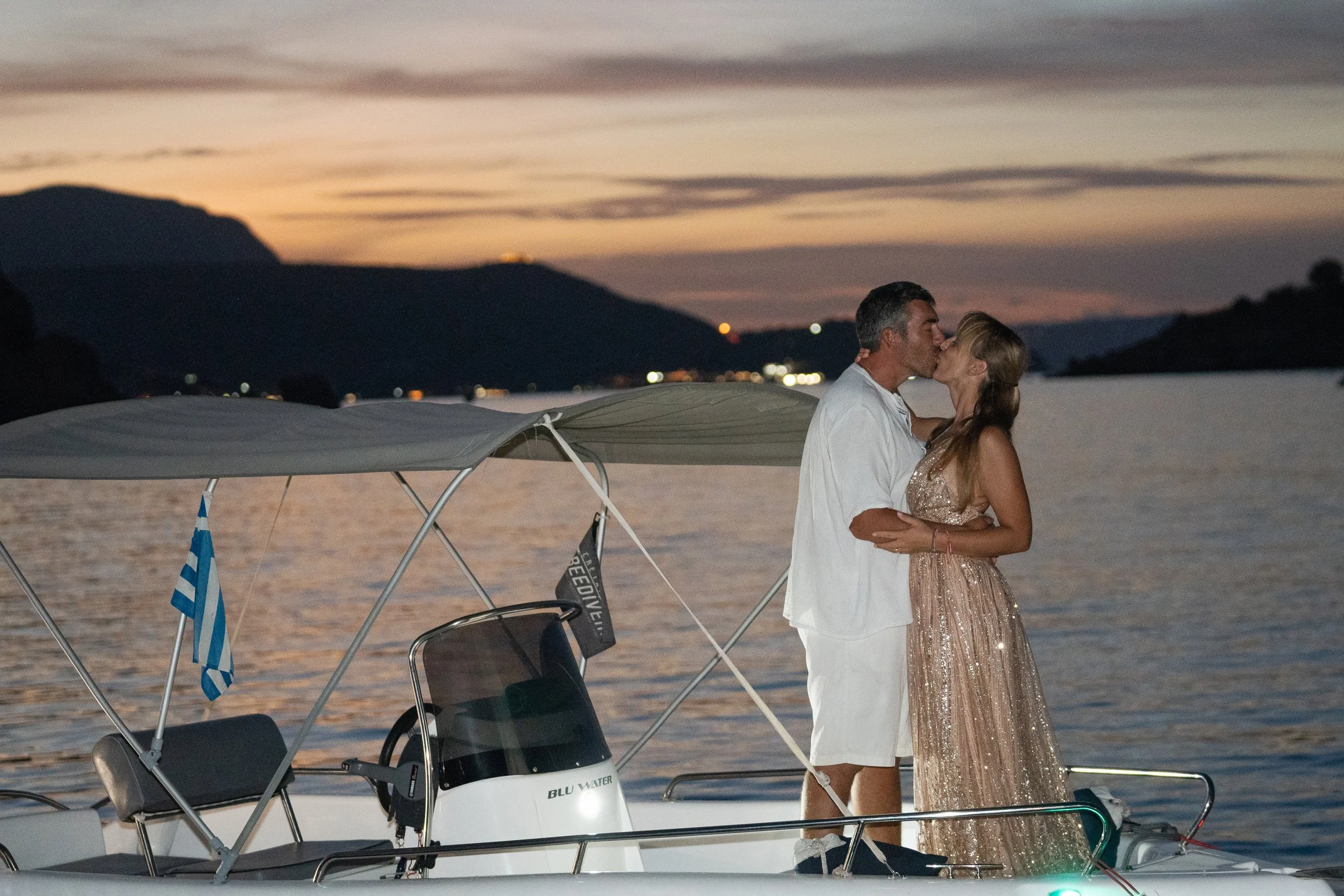 Couple kisses on a boat in Greece with a beautiful sunset behind them.