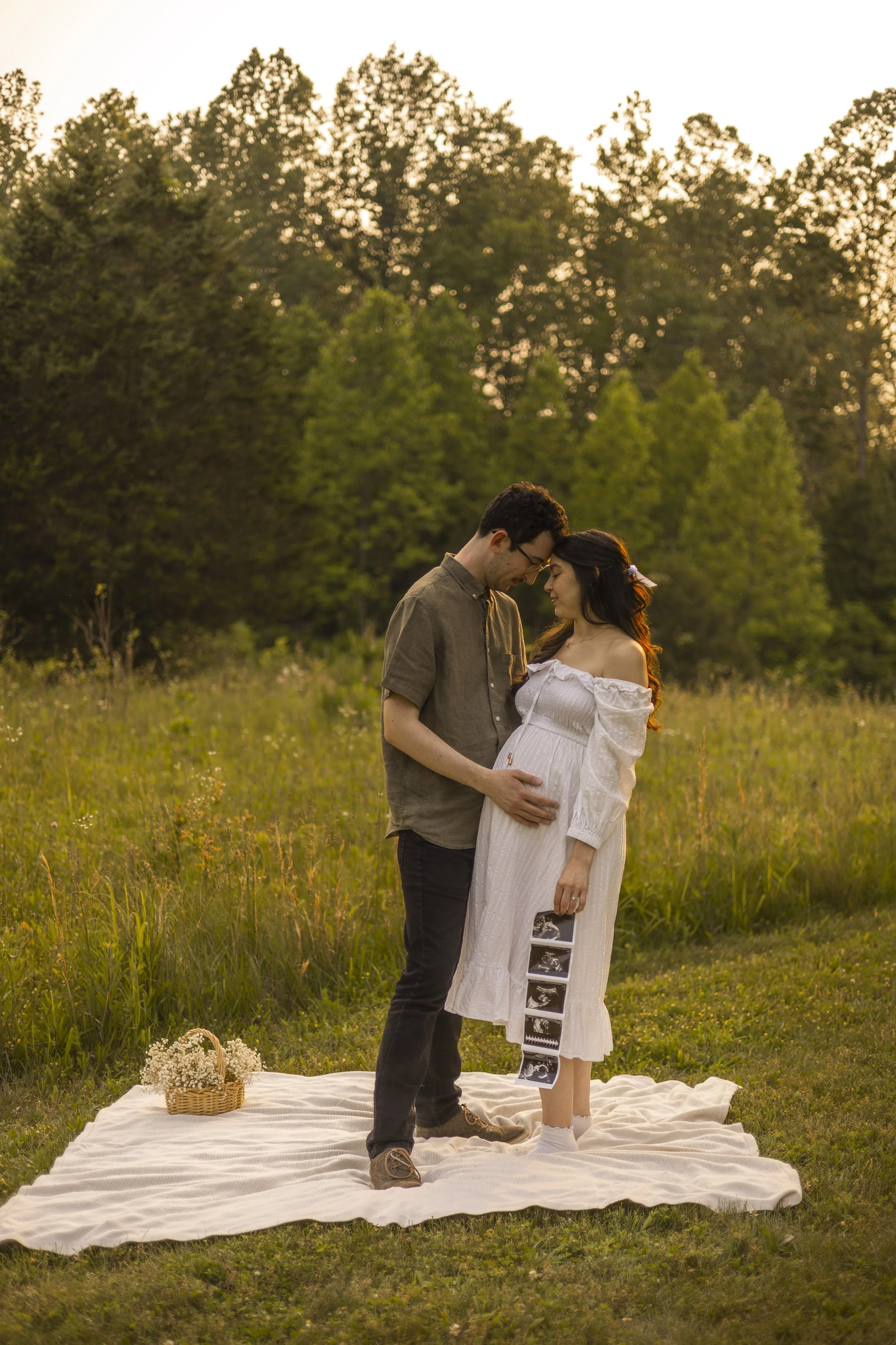 A pregnant woman and a man standing on a picnic blanket in a field at sunset, holding ultrasound images, and touching foreheads in a tender moment.
