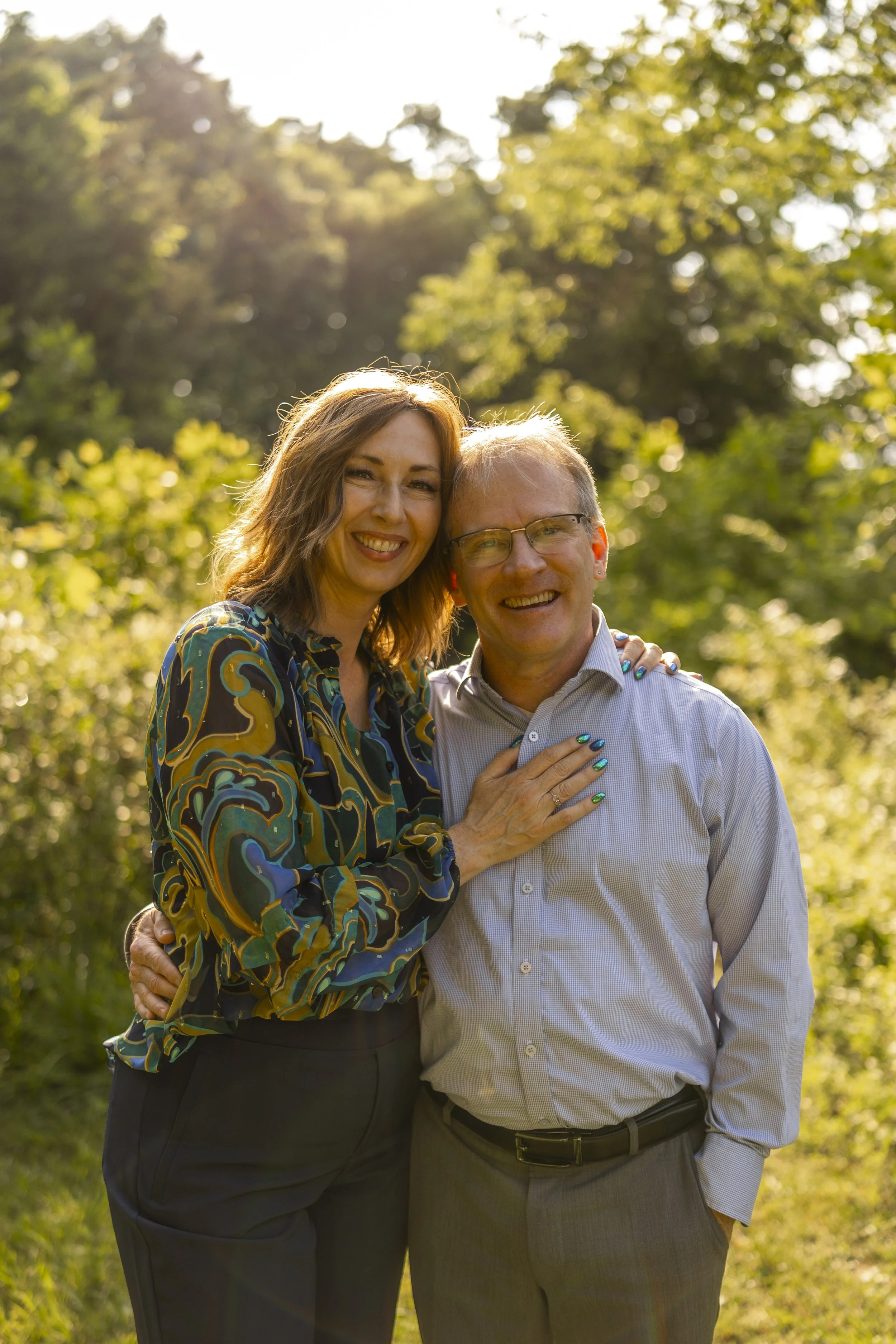 A woman and a man smiling outdoors in a sunny, green park, hugging and showing affection.