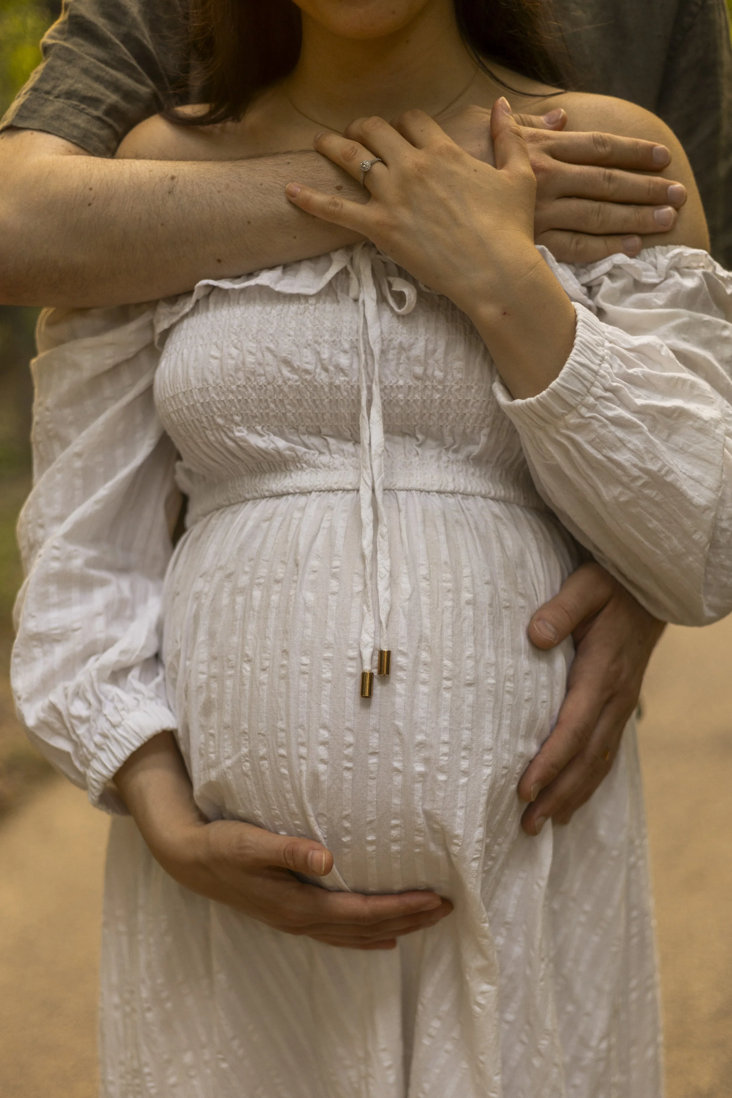 Close-up of a pregnant woman in a white dress being held by her partner, with one hand on her chest and the other on her belly. The woman's engagement ring is visible on her left hand.