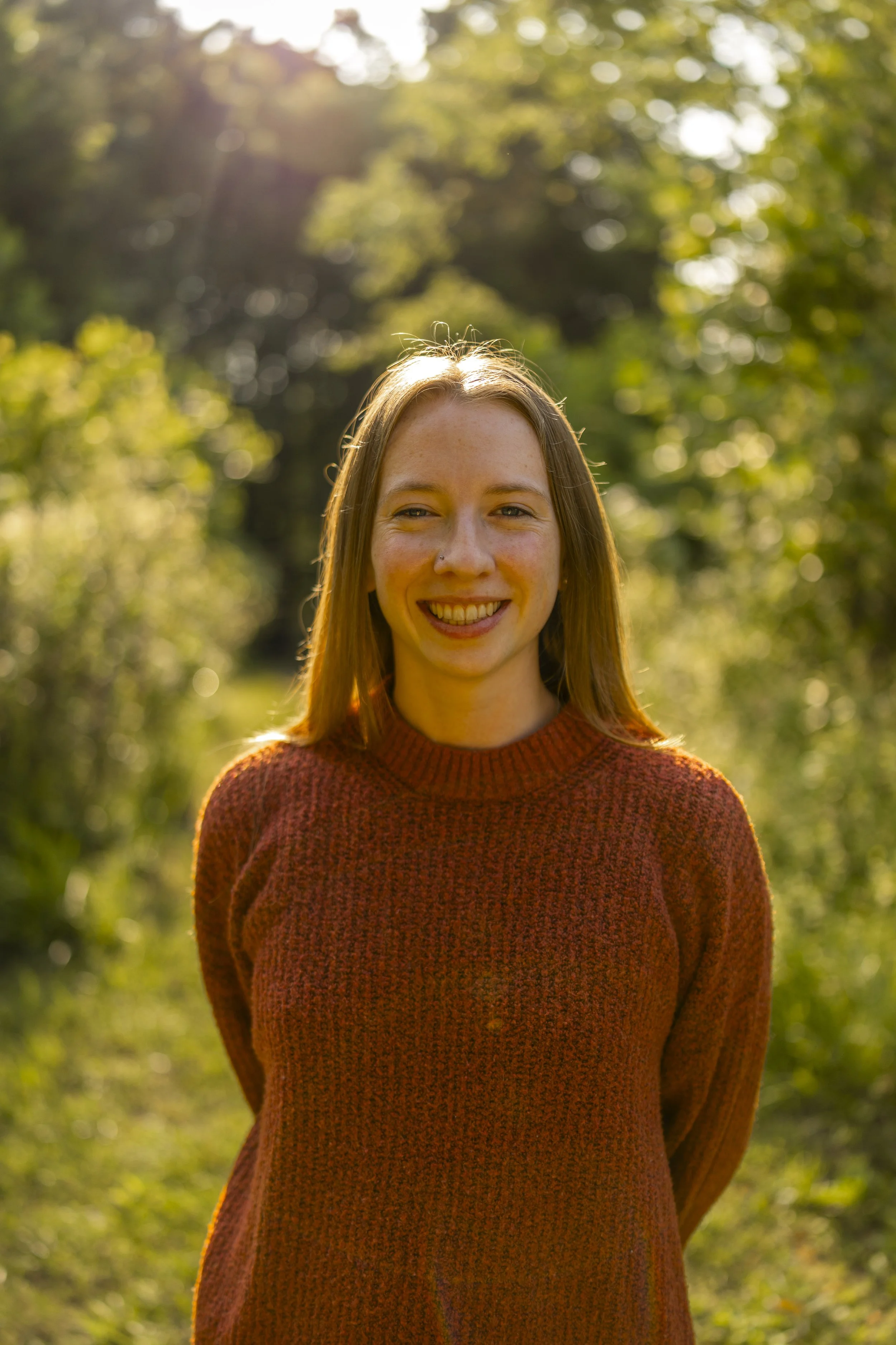 A young woman with long brown hair smiling outdoors during daytime, standing in front of green trees with sunlight filtering through.