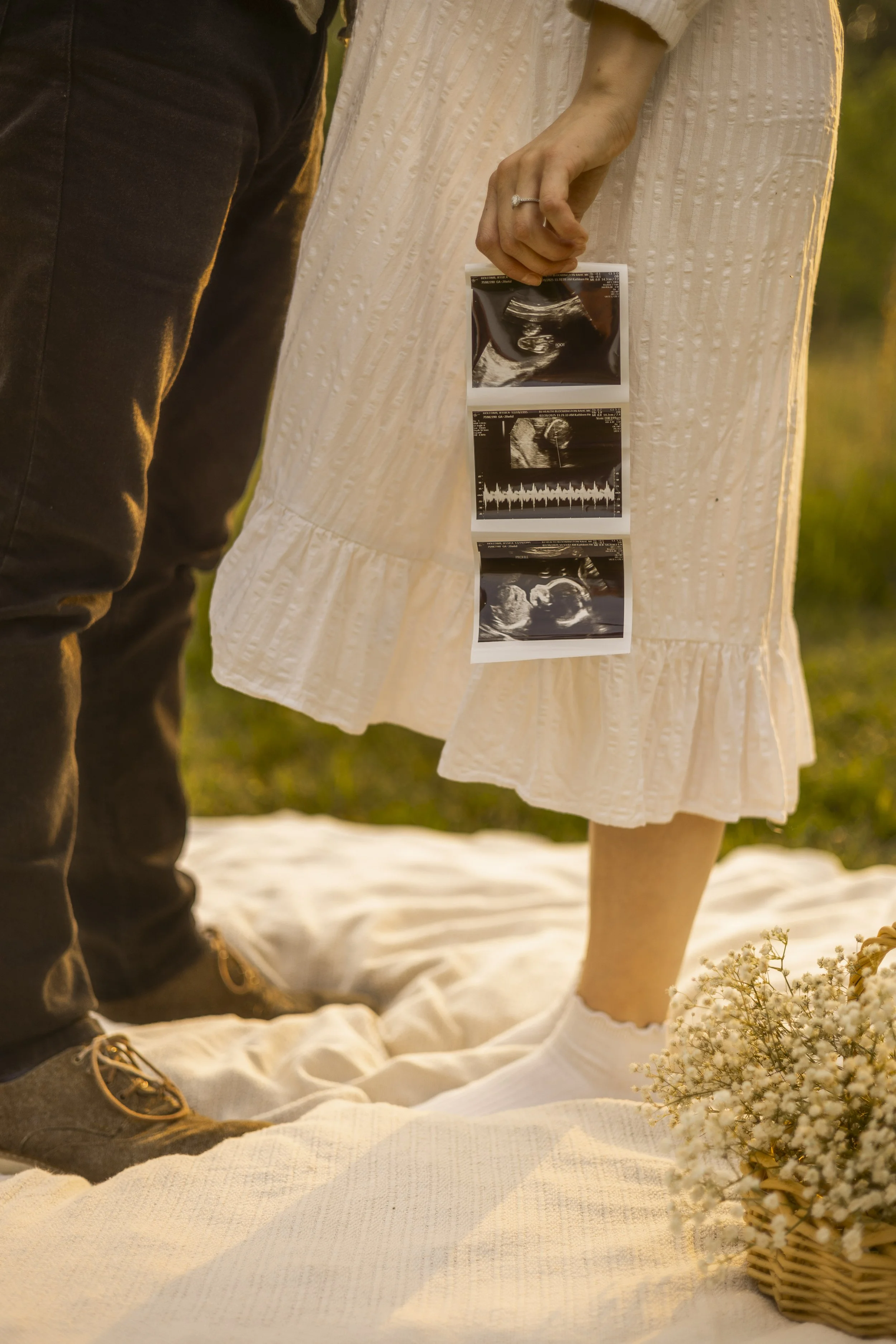 A pregnant woman holding ultrasound images and a man standing next to her in an outdoor setting with flowers and a blanket on the ground.