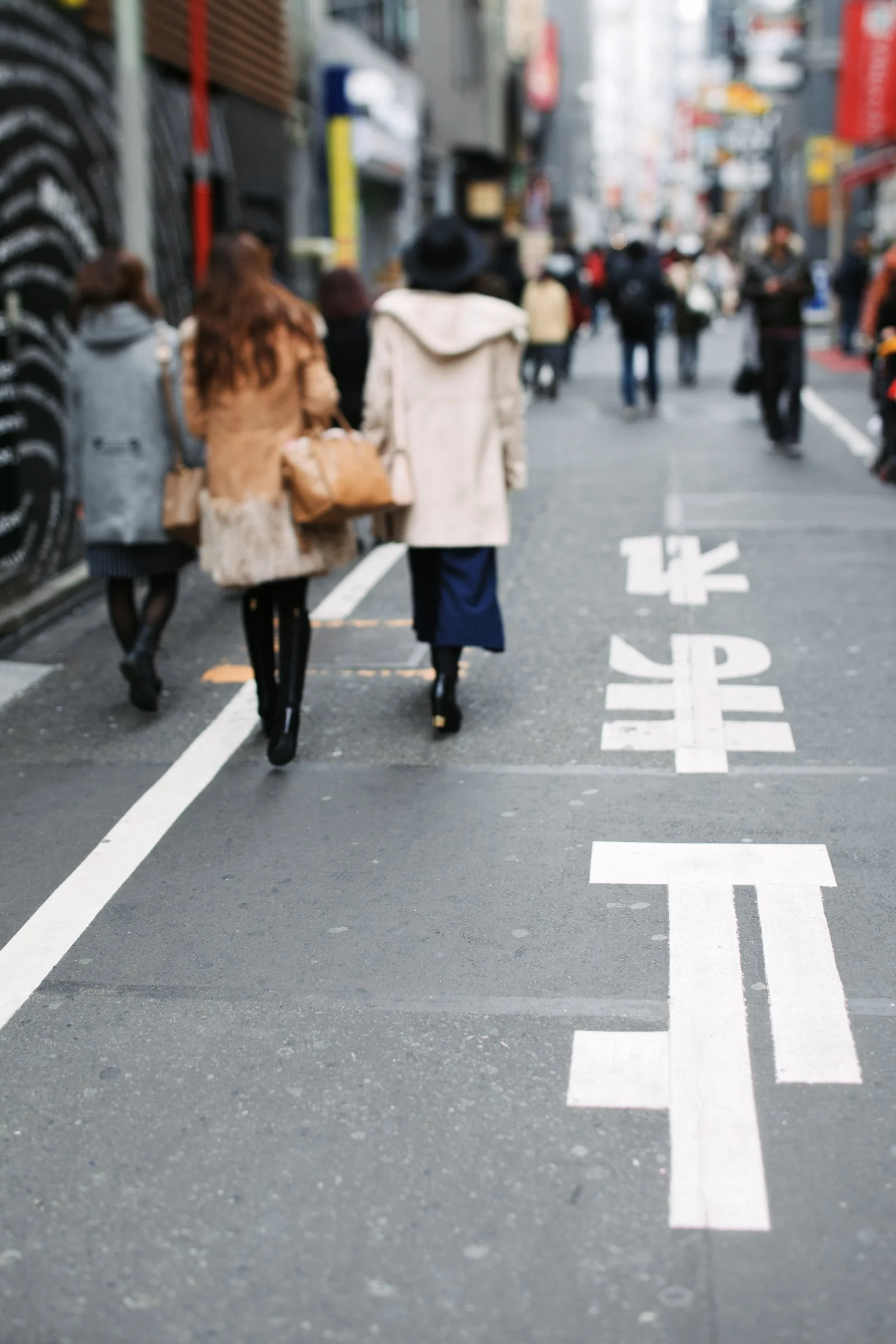 A busy city street with pedestrians walking, some wearing coats and hats. The street has clear white lane markings and signs, with buildings and advertisement banners visible in the background.