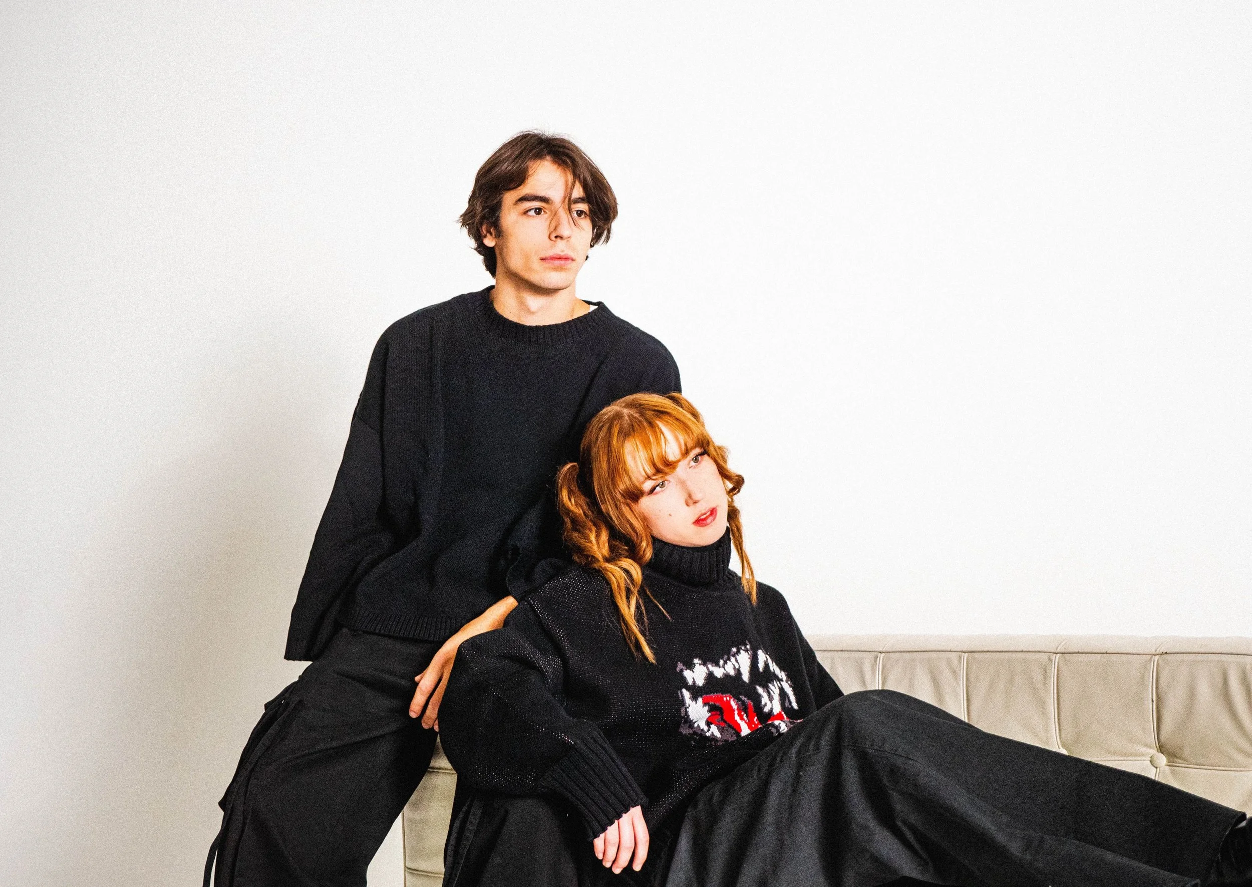 A young man with medium-length brown hair, wearing a black sweater, standing next to a young woman with red hair and glasses, sitting on a beige couch against a plain white wall.