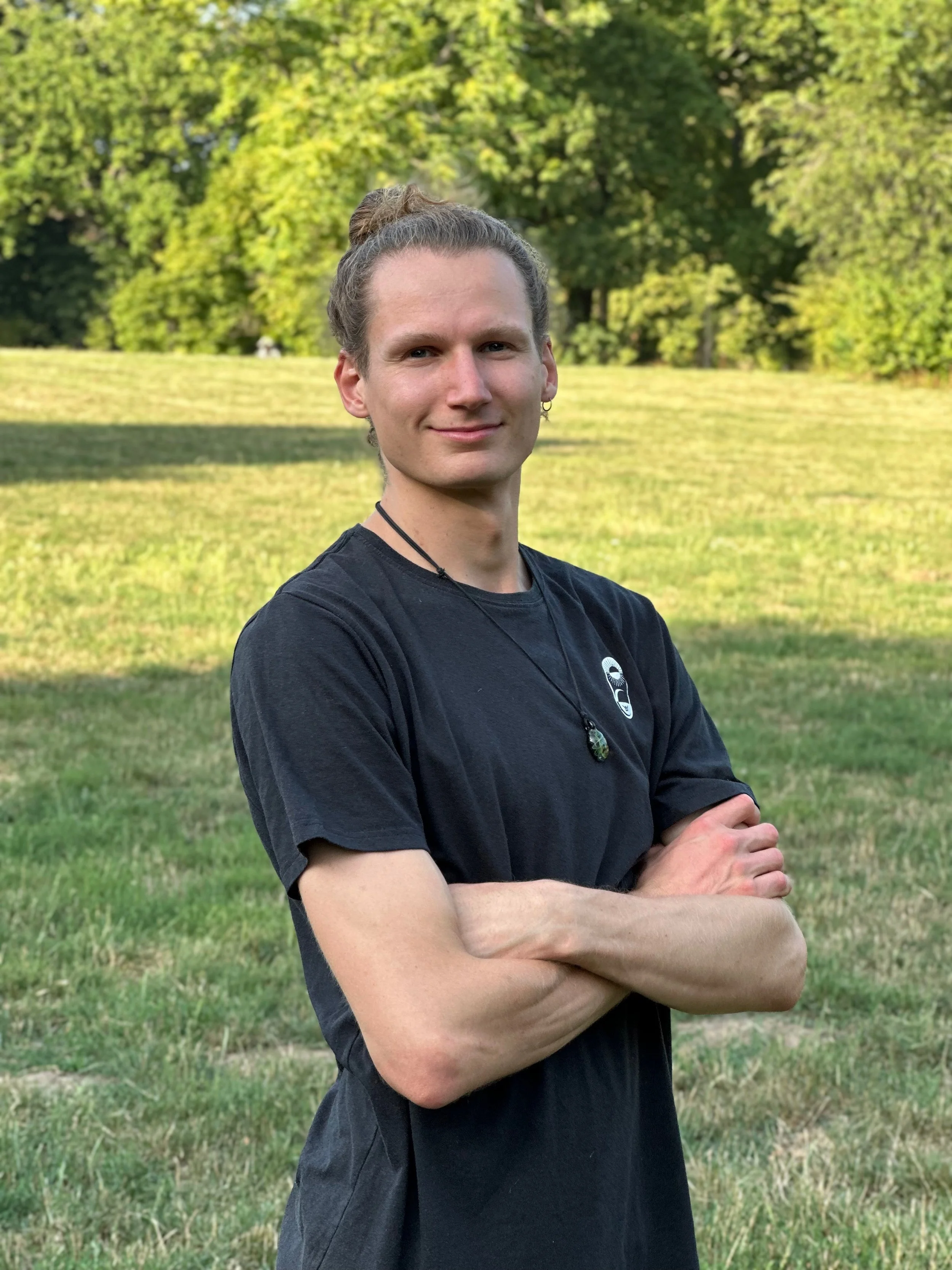 Finn König with long hair tied back, wearing a black t-shirt and jewelry, standing outdoors in a park with green trees and grass, arms crossed, smiling gently.
