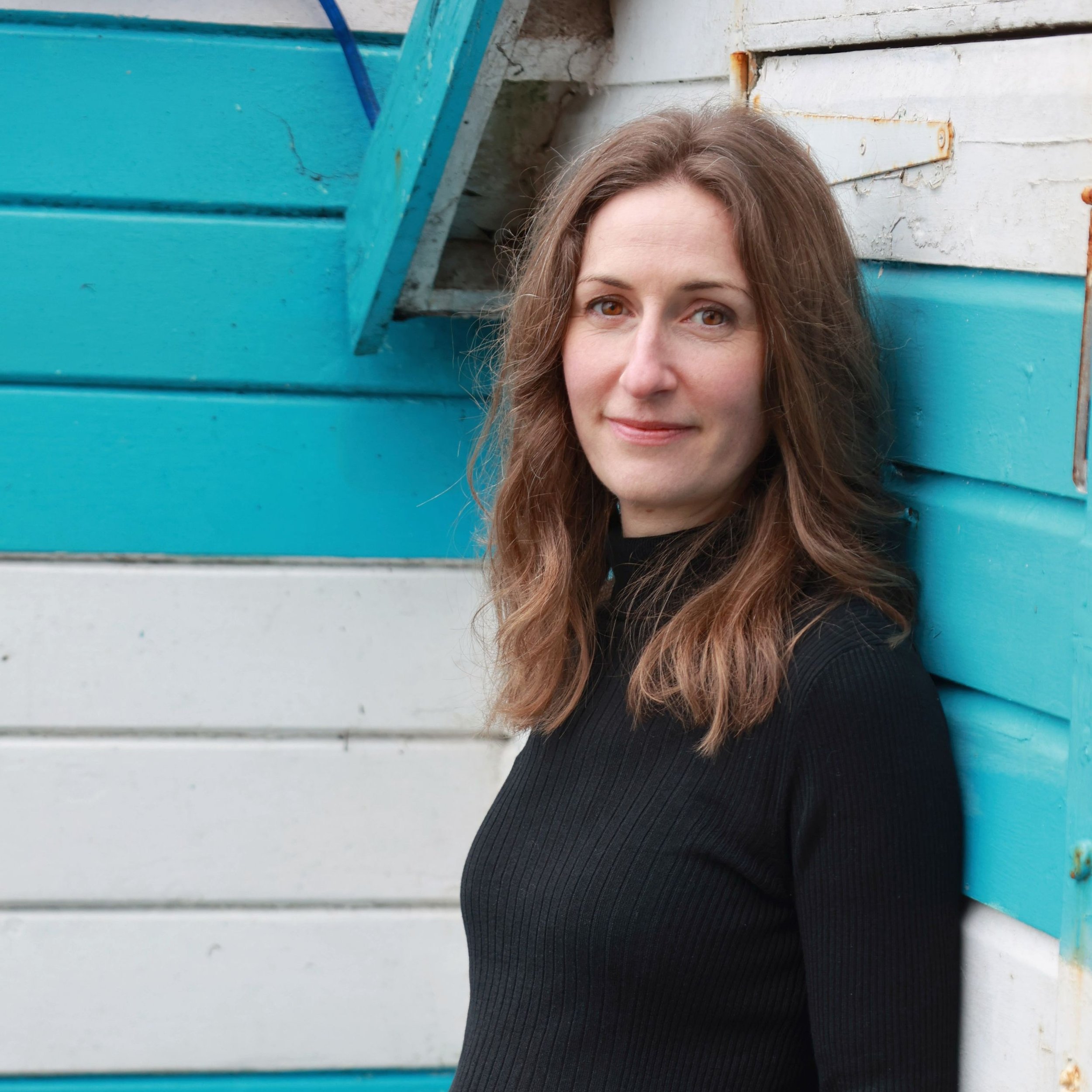 Close-up of Charlotte Horder with long brown hair and brown eyes smiling in front of a colorful textured wooden wall painted in shades of blue, white, and weathered wood.