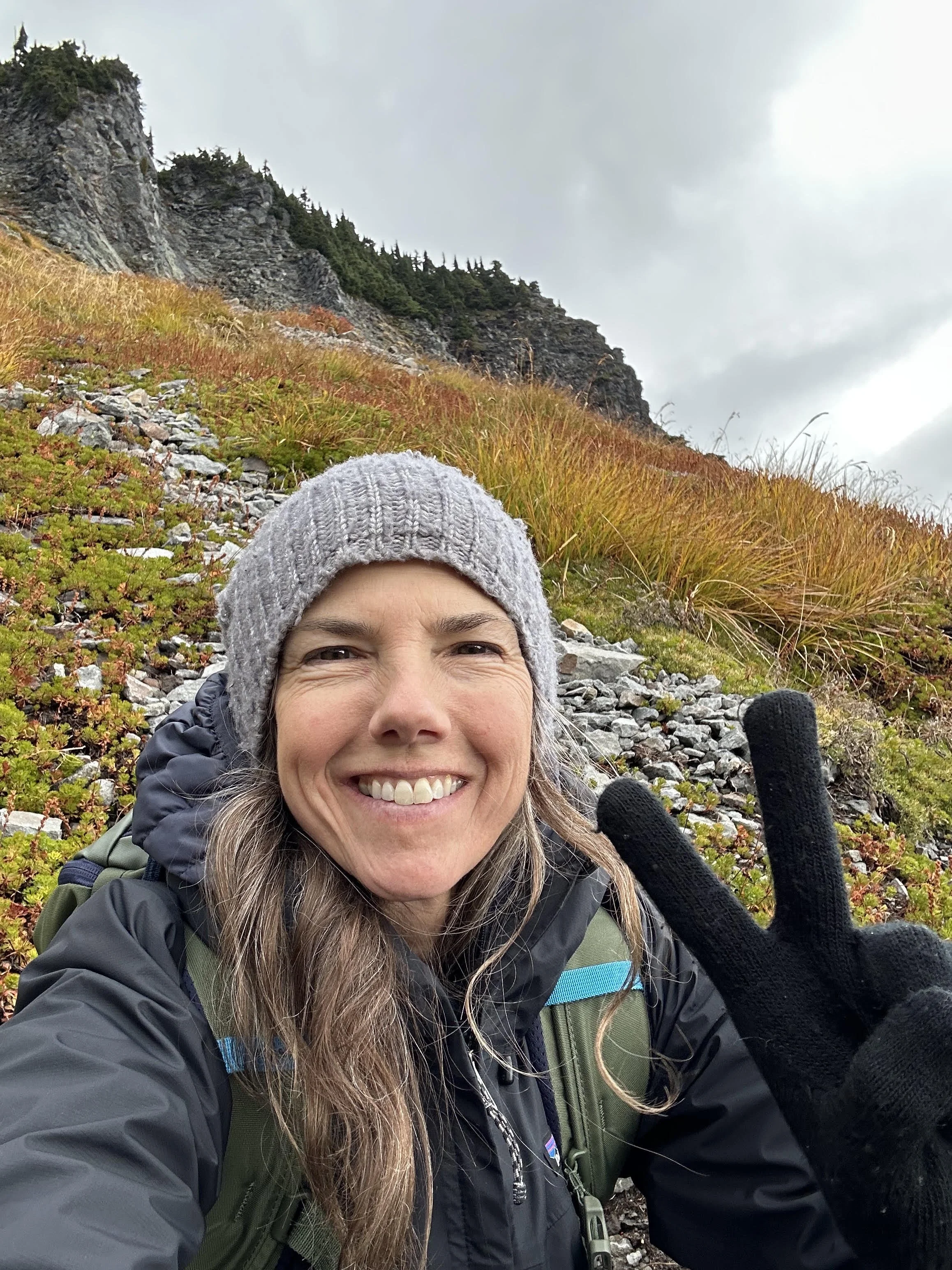 Kelly Isabelle Demarco taking a selfie outdoors during a hike on a mountain trail with rocky terrain and colorful plants, wearing a gray beanie and black waterproof jacket, showing a peace sign with her gloved hand, smiling under cloudy skies.
