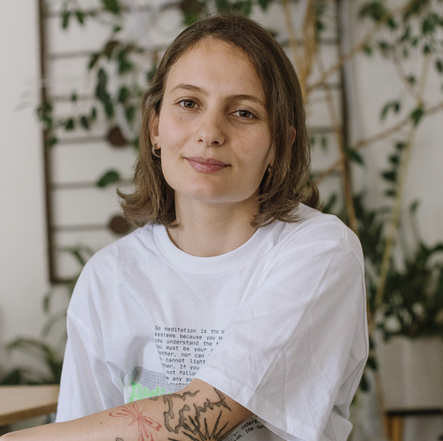 Juana Chiurchiu with shoulder-length brown hair, wearing a white t-shirt with text, standing outdoors with greenery in the background.
