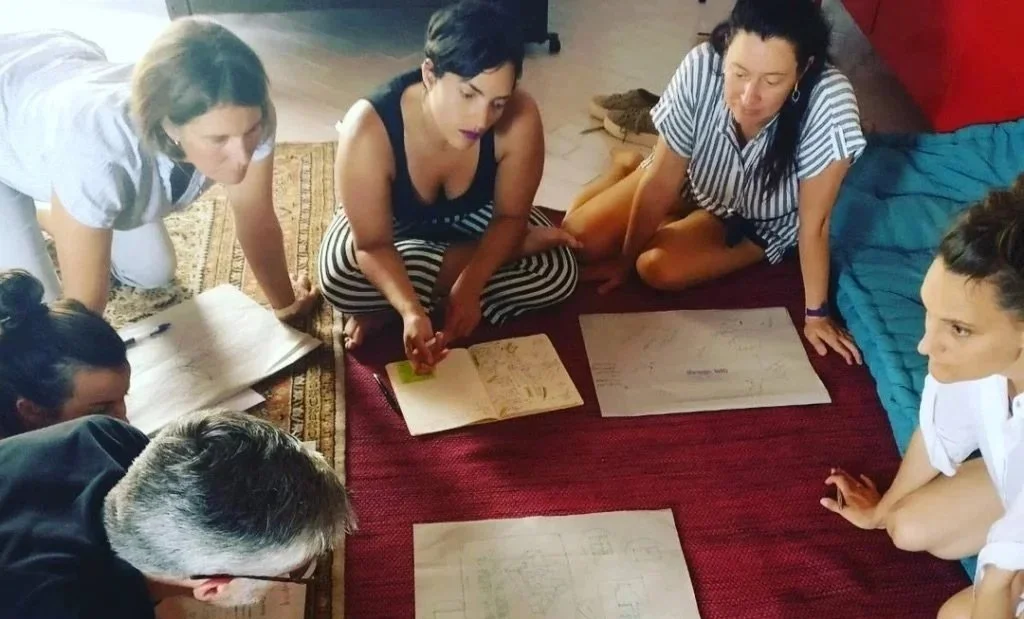 Group of six women sitting and kneeling on a red carpeted floor gathered around documents and a notebook, engaged in a discussion.