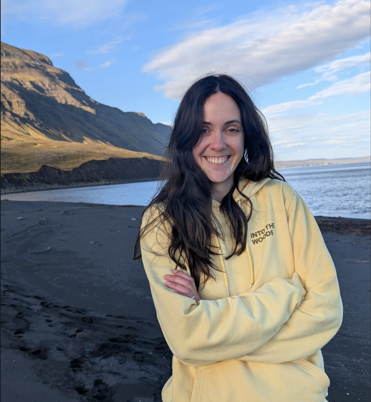 Constanza Reca with dark wavy hair wearing a yellow hoodie with the text "INTO THE WOODS" on it, standing on a black sand beach near a body of water with mountains and a partly cloudy sky in the background, smiling with arms crossed.