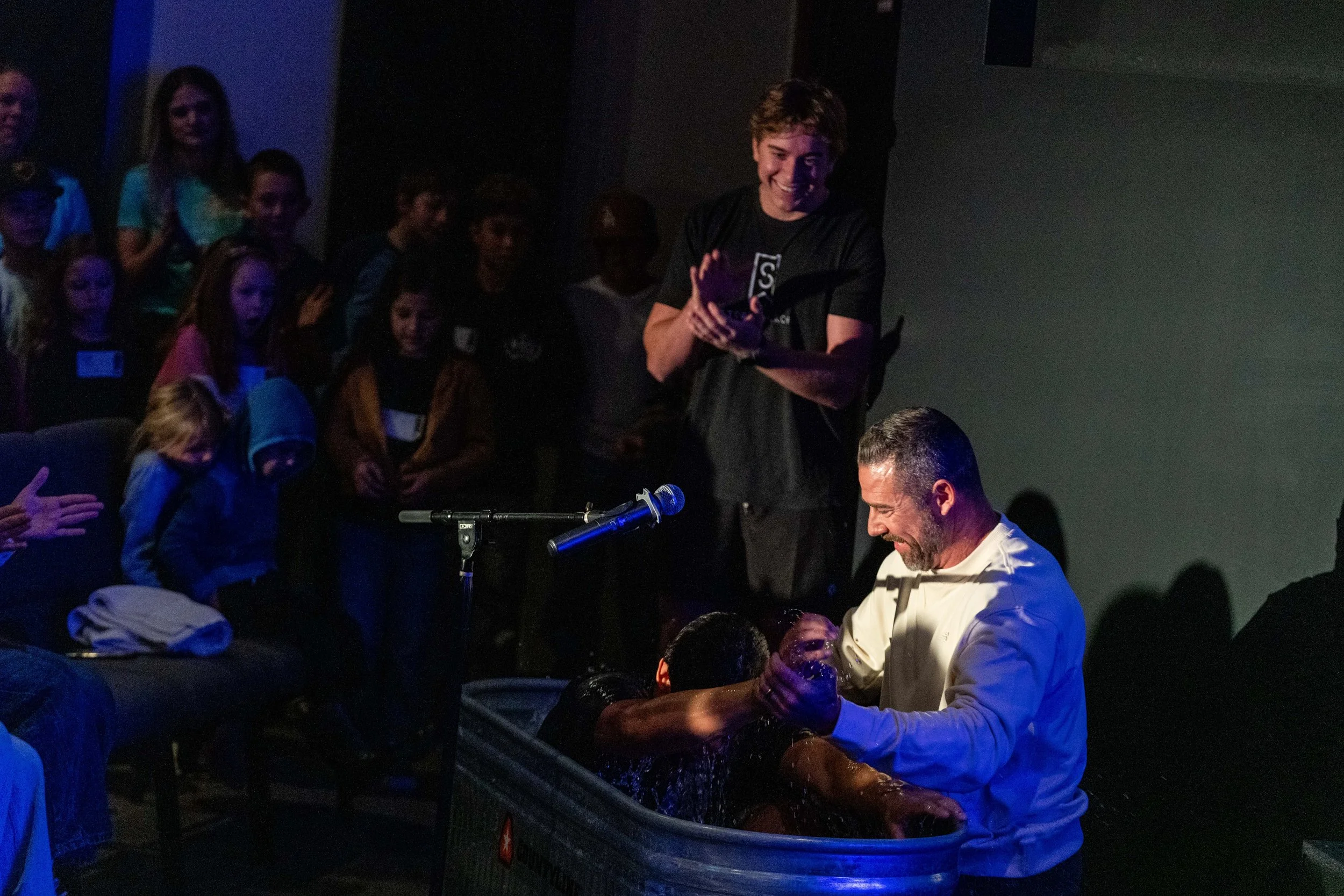 A man is baptizing a person in a small pool while a group of children and adults watch. The man is dressed in white, and the atmosphere appears joyful and celebratory.