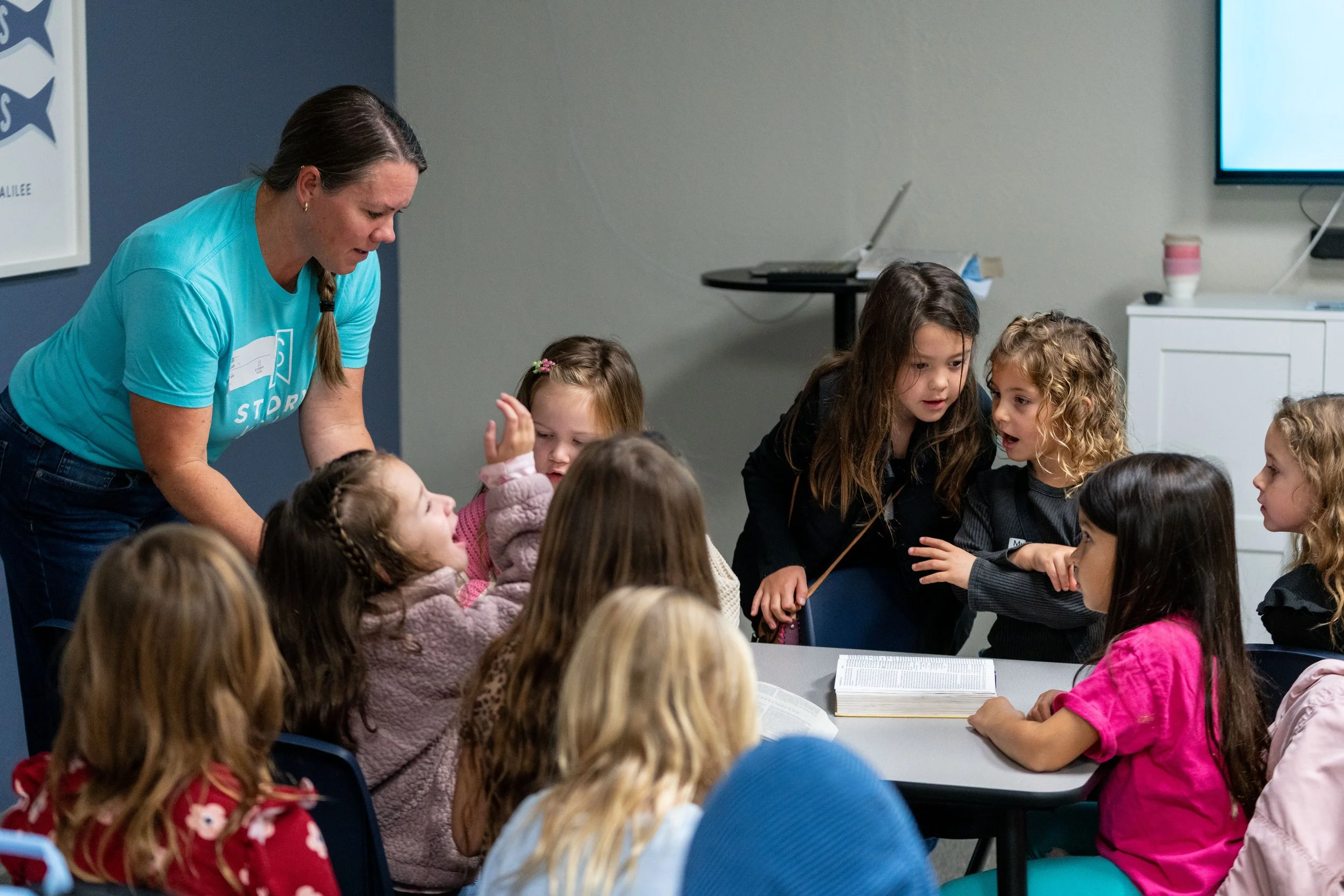 Group of young girls gathered around a table in a classroom, listening to a woman who is speaking to them.
