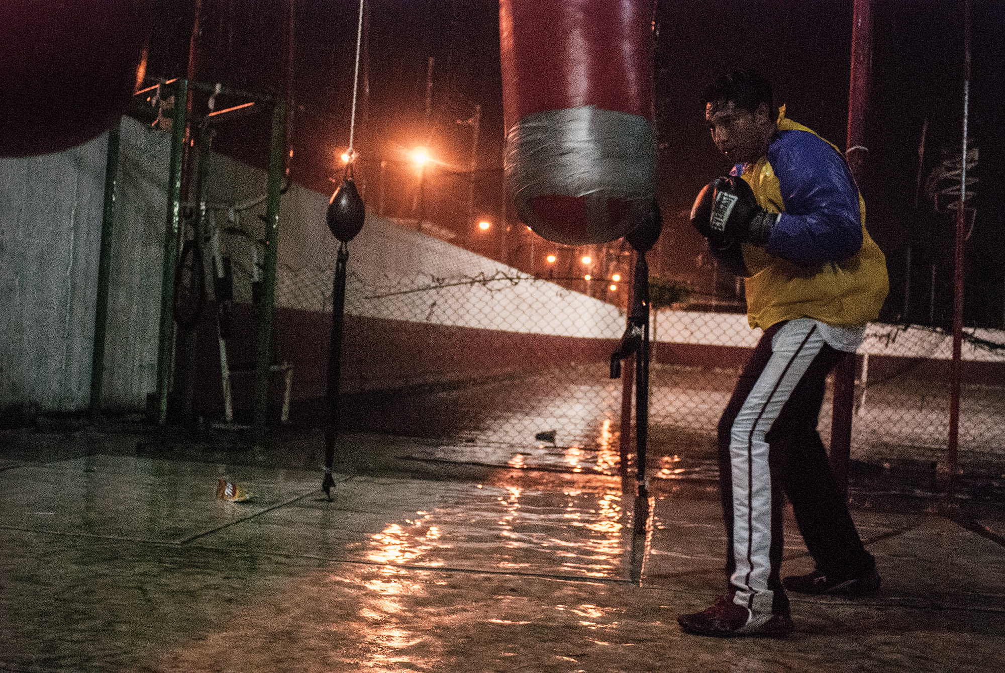 In the outdoor boxing gym of coach Lopez, a boxer works the punching bag on a rainy night.
