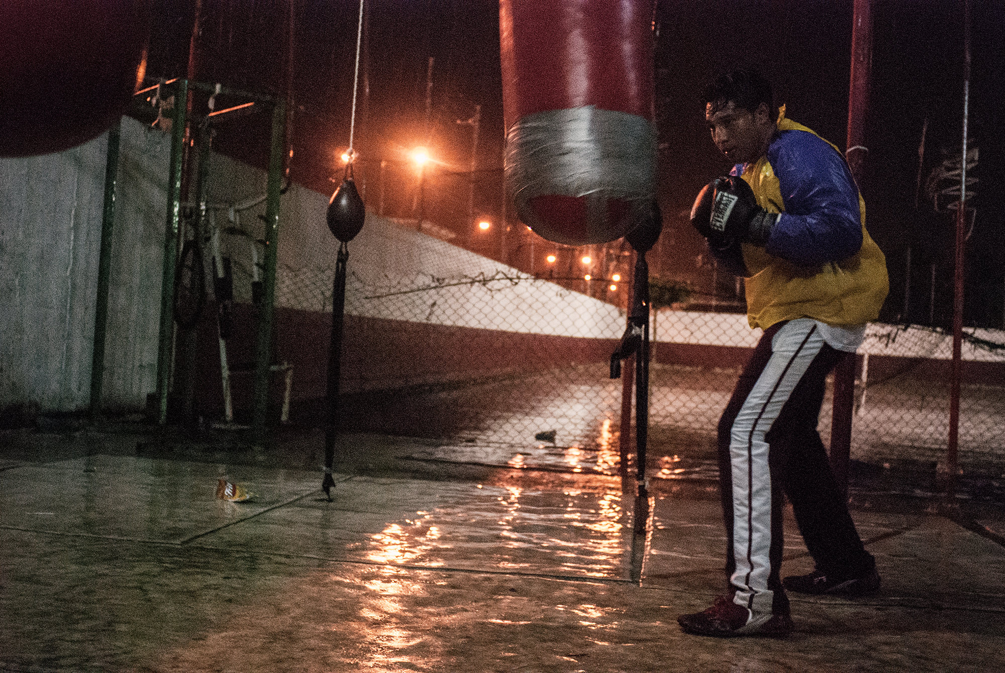  In the outdoor boxing gym of coach Lopez, a boxer works the punching bag on a rainy night. 