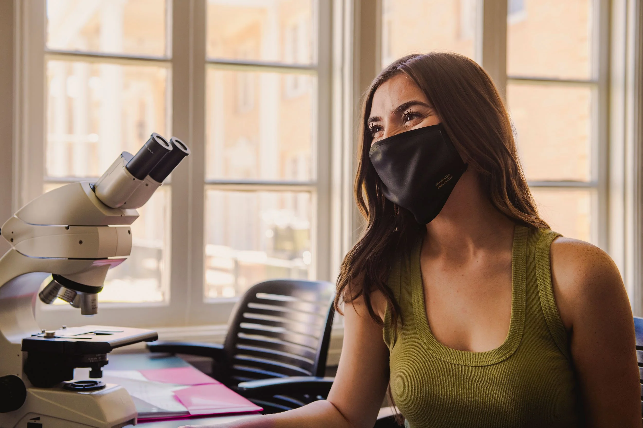 Woman wearing black face mask sitting by a microscope in a well-lit room for Arizona State University, captured by a Phoenix advertising photographer.